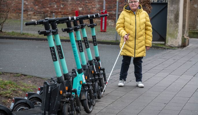 Eine Frau in gelber Jacke steht an einer Reihe abgestellter E-Roller. Sie orientiert sich mithilfe eines Langstocks.