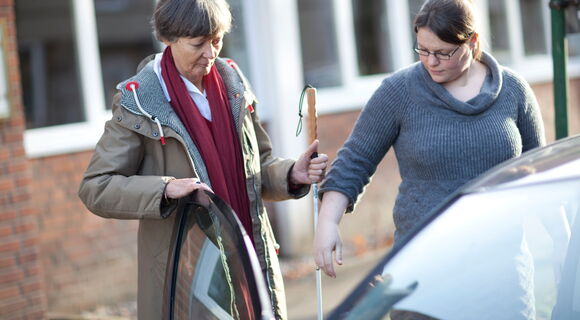 Zwei Frauen mittleren Alters stehen an einer offenen Autotür. Eine von ihnen hält der anderen ihren Arm hin. Diese hat einen Langstock dabei und hält sich mit einer Hand an der Autotüre.