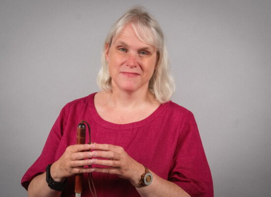 Porträt von Ute Lutzenberger. Blonde, glatte Haare fallen bis auf ihre Schultern. Vor der Brust hat sie die Fingerspitzen aneinander gelegt und hält ihren weißen Langstock. Sie trägt ein luftiges, rotes Oberteil und eine dunkle Uhr am Handgelenk.