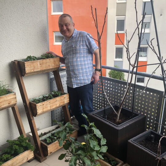 Rainer Heim trägt ein helles, kariertes Hemd und steht lächelnd auf seinem Balkon. Neben ihm ein dreistufiges Hochbeet mit Kräutern, vor ihm einige Blumenkübel mit wachsenden Pflanzen.