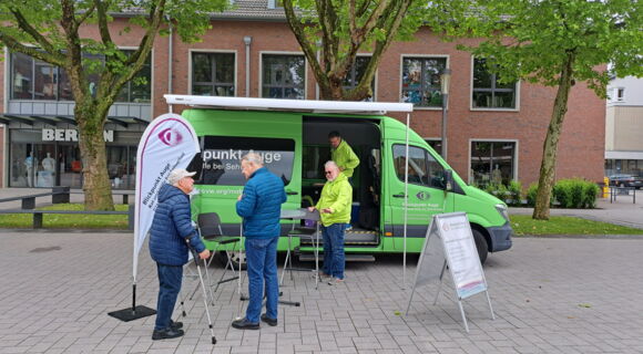 Das Beratungsmobil des Blinden- und Sehbehindertenvereins Westfalen ist   auf einem Platz geparkt. Johannes Willenberg steht vor dem Wagen an einem  Stehtisch und blickt zu zwei älteren Herren, die sich dem Fahrzeug nähern.