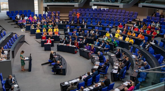 Das Bild zeigt den Plenarsaal des Bundestags von schräg oben. Die Abgeordneten sitzen im Halbrund um die Regierungsbank und das Rednerpult. Am Rednerpult steht eine junge Frau mit Pferdeschwanz in einem Sommerkleid mit floralen Elementen und Sandalen.