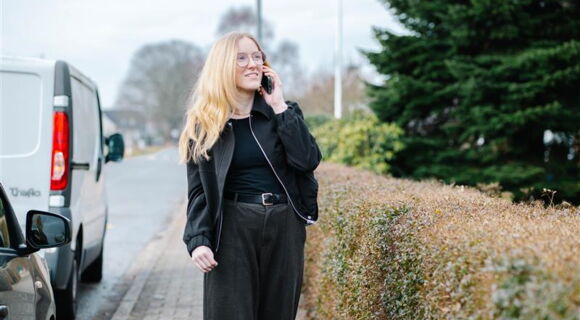 Claire Horsbrugh steht telefonierend an einer Straße. Sie hat langes blondes Haar und trägt eine schwarze Jacke und ein schwarzes Oberteil. Im Hintergrund sind Bäume und Büsche zu sehen.