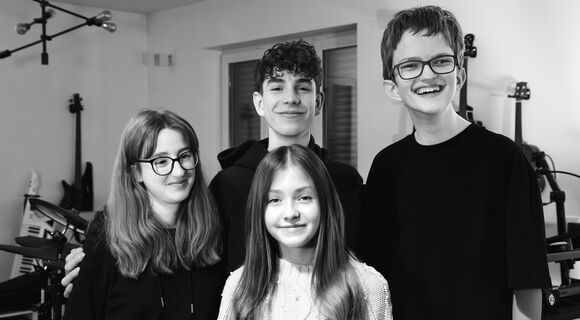 Die Band Rockytrio, vier Jugendliche zwischen 13 und 16, stehen in einem Musikraum vor Instrumenten und schauen freundlich lächelnd in die Kamera.