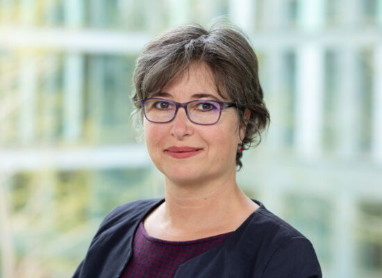 Porträt von Julia Gebrande: Eine Frau mit kurzem braunem Haar und Brille blickt in die Kamera. Sie trägt ein dunkles Jackett über einem gemusterten Oberteil. Der Hintergrund ist hell und unscharf.