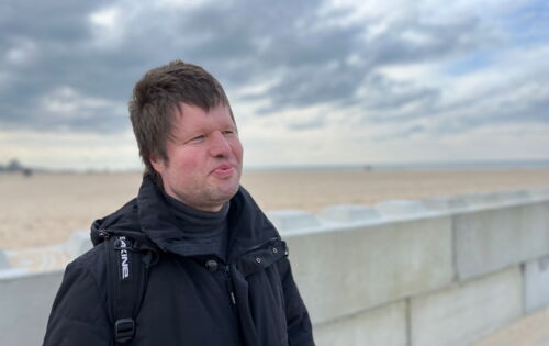 Marco Mers in dunkler Jacke an einem weitläufigen Ostseestrand. Er hat kurzes Haar und lächelt. Der Himmel ist wolkenverhangen.
