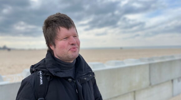 Marco Mers in dunkler Jacke an einem weitläufigen Ostseestrand. Er hat kurzes Haar und lächelt. Der Himmel ist wolkenverhangen.