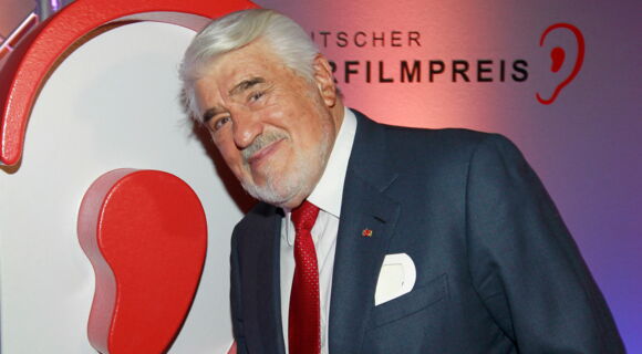 Mario Adorf, ein älterer grauhaariger Mann, steht lächelnd vor einem Plakat mit der Aufschrift "Deutscher Hörfilmpreis". Er trägt Anzug und Krawatte.