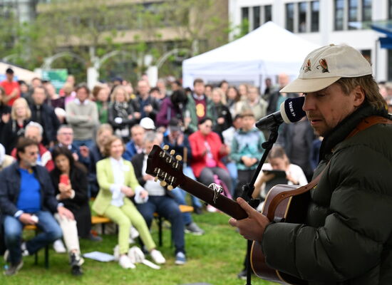 Ein Blick von der Außentribüne auf dem Berliner Platz ins Publikum der Eröffnungsfeier: rechts im Bildvordergrund steht scharf zu sehen Philipp Poisel mit seiner Gitarre, den leicht verschwommenen Besucherinnen und Besuchern zugewandt.