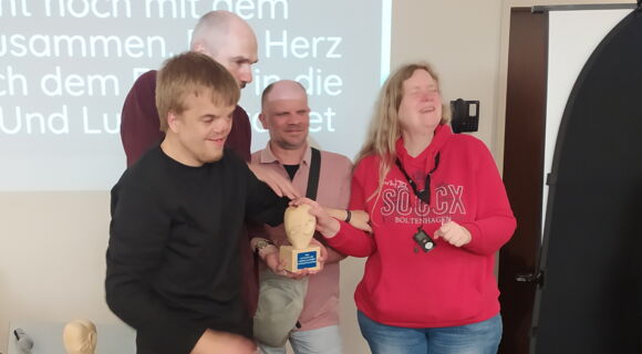 Bei der Preisverleihung nehmen Lucas Brinse, Reiner Delgado und Annette Sander den Preis entgegen. Sie halten die Skulptur gemeinsam in den Händen: Das Herz aus hellem Holz, auf dem ein Auge und ein Ohr zu sehen sind, steht auf einem Sockel.