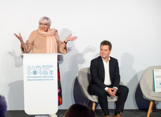 Claudia Roth, Ministerin für Kultur und Medien, steht mit ausgebreiteten Armen an einem Rednerpult, darauf ein Logo mit dem Text: „Kultur braucht Inklusion, Inklusion braucht Kultur“. Daneben sitzt der Bundesbehindertenbeauftragte, Jürgen Dusel.