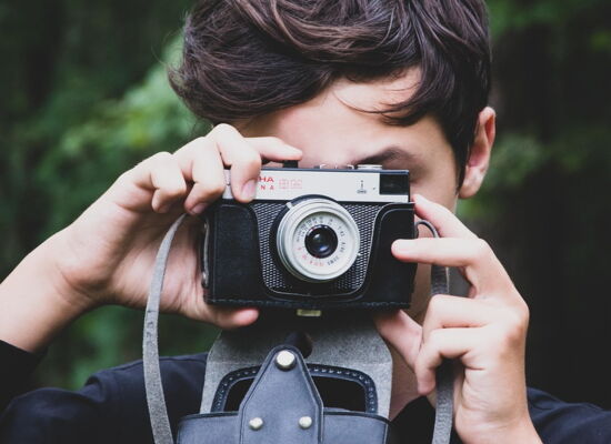 Ein junger Mensch mit dunklen Haaren hält eine analoge Kamera vor sein Gesicht und fotografiert. Die Kamera hat ein Vintage-Design mit schwarzem Gehäuse und silbernen Details. Der Hintergrund ist grün und unscharf.