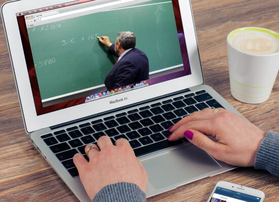 Eine Person tippt auf einem MacBook, auf dessen Bildschirm ein Video mit einem Lehrer läuft, der an eine grüne Tafel schreibt. Die Person trägt graue Kleidung, hat lila lackierte Nägel und trägt Ringe. Daneben stehen eine Tasse Kaffee und ein Smartpho
