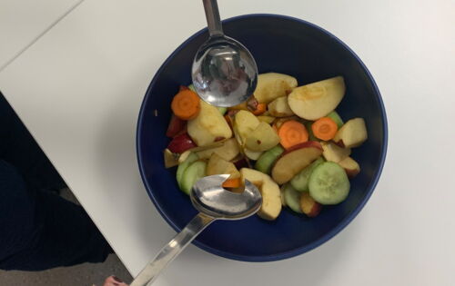 Ein bunter Salat wird in einer dunklen Schüssel mit Salatbesteck vermengt. Das Gefäß hebt sich kontrastreich von der hellen Arbeitsplatte ab.