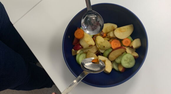 Ein bunter Salat wird in einer dunklen Schüssel mit Salatbesteck vermengt. Das Gefäß hebt sich kontrastreich von der hellen Arbeitsplatte ab.