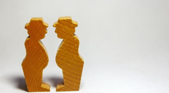 Auf einem weißen Hintergrund stehen sich zwei kleine Holzfiguren gegenüber. Sie sehen aus wie zwei untersetzte Männer mit Hut.