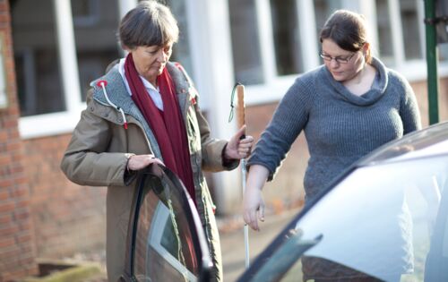 Zwei Frauen mittleren Alters stehen an einer offenen Autotür. Eine von ihnen hält der anderen ihren Arm hin. Diese hat einen Langstock dabei und hält sich mit einer Hand an der Autotüre.