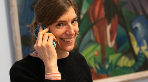 Eine Frau mit glatten, braunen Haaren hält sich lächelnd ein Smartphone ans Ohr. Hinter ihr ist ein abstraktes Gemälde zu erkennen.