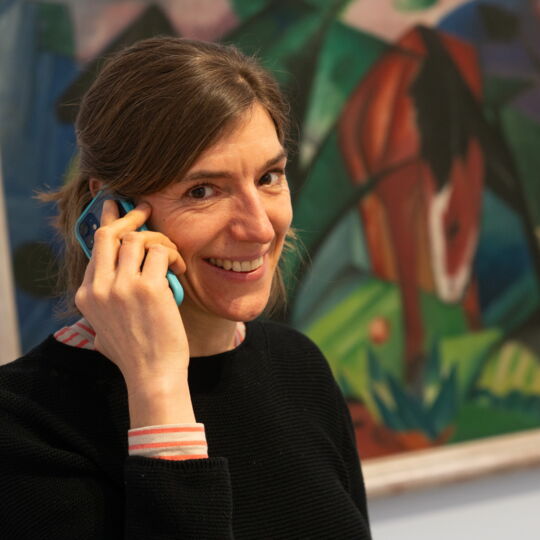 Eine Frau mit glatten, braunen Haaren hält sich lächelnd ein Smartphone ans Ohr. Hinter ihr ist ein abstraktes Gemälde zu erkennen.