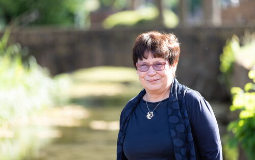 Margit Giegerich trägt kurzes dunkles Haar, eine Brille, dunkle Kleidung und eine Kette mit blütenförmigem Anhänger. Sie steht auf einem sonnigen Weg, im Hintergrund sind verschwommen Büsche und Bäume zu sehen.