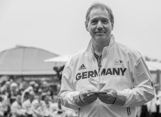 Dr. Karl Quade mit einer Trophäe bei den Paralympics in Rio de Janeiro. Er trägt kurzes, schütteres Haar und hat einen Dreitagebart. Er lächelt in die Kamera. Auf seiner hellen Jacke steht „GERMANY“.