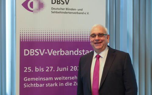 DBSV-Präsident Hans-Werner Lange steht lächelnd vor einem Plakat, das auf den Verbandstag 2026 hinweist. Er hat kurzes graues Haar, trägt eine getönte Brille, Anzug und Krawatte.