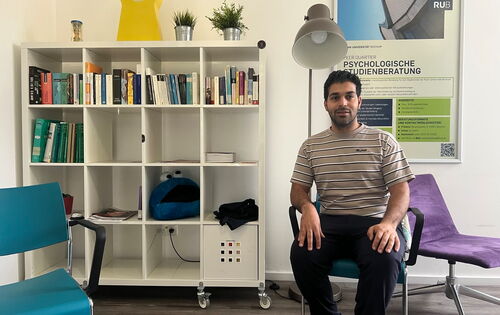 Saiman Mehra sitzt im Raum der Psychologischen Studienberatung. Er hat dunkles Haar und einen Vollbart. Hinter ihm an der Wand steht ein weißes Regal mit quadratischen Fächern, gefüllt mit Büchern und anderen Dingen.