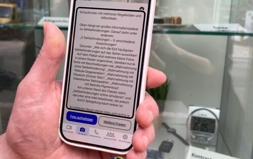 Eine linke Hand richtet ein Smartphone auf ein Schaufenster. Auf dem Display ist viel Text zu erkennen