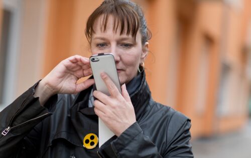 Eine Frau mit Pony-Haarschnitt schaut konzentriert auf ihr Smartphone. An ihrer schwarzen Jacke befindet sich ein gelber Anstecker mit drei Punkten.