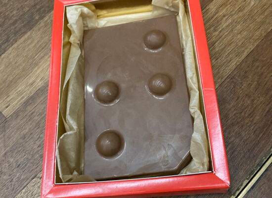 In einer roten Schachtel liegt eine Tafel Schokolade. Darauf ist der Buchstabe "T" in Braille abgedruckt.