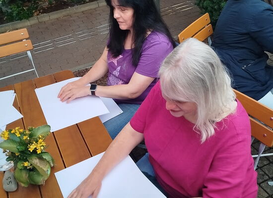 Zwei Frauen sitzen an einem Holztisch und studieren mit den Fingern je eine Wanderkarte in Braille. Die Frau links ist Yvonne Bühr. Sie hat langes dunkles Haar.