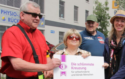 Auf einem Gehweg: Bodo Rinas (links) und Margrit Richter (2.v.l.) übergeben die plakatgroße Urkunde zur schlimmsten Kreuzung an die Behindertenbeauftragte von Königs Wusterhausen, Friederike Weigelt (mittig). Weitere Beteiligte schauen zu.
