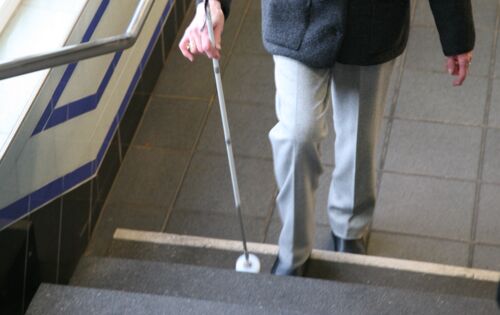 Eine ältere Frau mit Langstock steigt eine Treppe hinauf. Die unterste Stufe ist deutlich visuell gekennzeichnet.