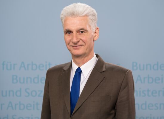 Rolf Schmachtenberg hat weißes, zurückgekämmtes Haar. Er trägt einen braunen Anzug, ein weißes Hemd und eine blaue Krawatte. Im Hintergrund ist unscharf der Text: „Bundesministerium für Arbeit und Soziales“ angedeutet.