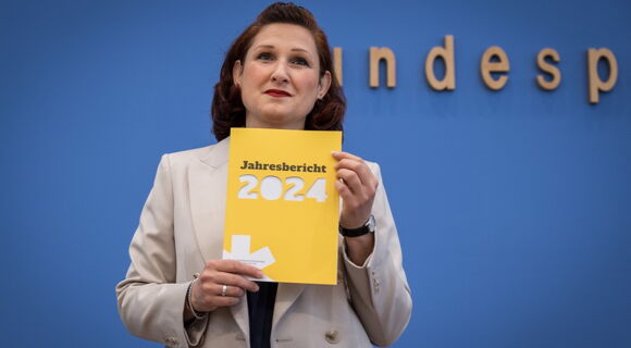 Das gelbe Heft mit dem Jahresbericht der Antidiskriminierungsstelle hält Ferda Ataman bei der Bundespressekonerenz hoch. Die unabhängigen Bundesbeauftragte hat braune, ondulierte Haare und trägt einen hellen Blazer.