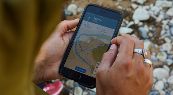 Jemand steht draußen auf Steinen und hält ein Handy mit der Navigations-App Komoot in der Hand. Darauf ist eine Route zu sehen. Der Mensch hat silberne Ringe und trägt eine grüne Jacke.