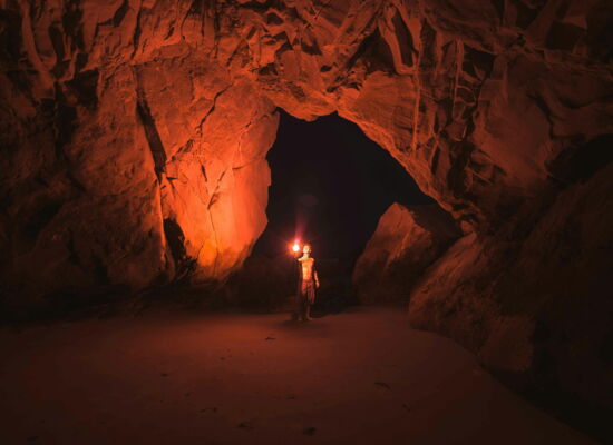 Ein junger Mann steht in einer Höhle. In seiner Hand hält er einen Stab mit einem hellen Licht, das die Wände der Höhle in warmes Licht taucht.