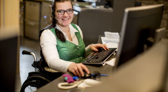Eine Frau mit breitem Lächeln sitzt in einem Büro am Computer. Sie nutzt einen Rollstuhl. Sie hat eine grüne Weste mit Knöpfen an, trägt eine Brille und hat ein Headset auf dem Kopf.