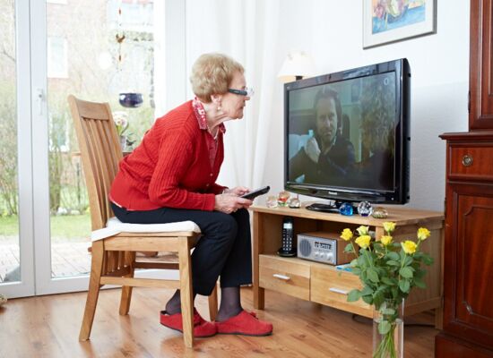 Eine ältere Frau in roter Strickjacke sitzt auf einem Stuhl direkt vor einem eingeschalteten Fernseher. In der Hand hält sie die Fernbedienung und beugt sich in Richtung des Bildschirms.