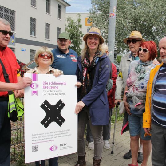 Auf einem Gehweg: Bodo Rinas (links) und Margrit Richter (2.v.l.) übergeben die plakatgroße Urkunde zur schlimmsten Kreuzung an die Behindertenbeauftragte von Königs Wusterhausen, Friederike Weigelt (mittig). Weitere Beteiligte schauen zu.