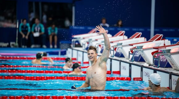 Taliso Engel winkt mit einem breiten Lachen aus dem Wasser in einem Schwimmbad. Er ist ein sehr muskulöser junger Mann. Neben ihm sind andere Schwimmer mit Badekappen.