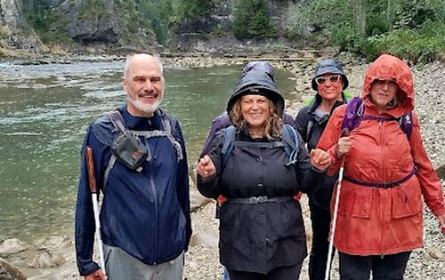 Reiner Delgado (links) und vier weitere Personen der Wandergruppe stehen lachend an einem steinigen Ufer. Sie tragen Outdoor-Jacken, Rucksäcke, Wanderschuhe und teilweise einen Langstock.