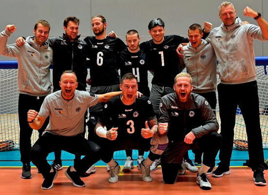Das Goalball-Team Deutschland steht jubelnd vor dem Tor. Es sind zehn weiße Männer in Sportkleidung. Sie haben einander die Arme über die Schulter gelegt