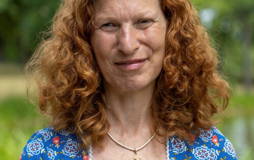 Brigitte Buchsein hat langes lockiges helles Haar. Sie trägt ein Shirt mit Blumen und eine Kette mit einem kleinen Kreuzanhänger.