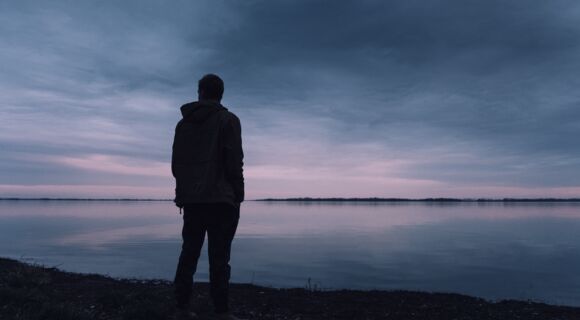 Die Silhouette einer männlich anmutenden Person, die alleine auf ein weites Gewässer blickt. Das Bild ist in dunklen Farben gehalten.