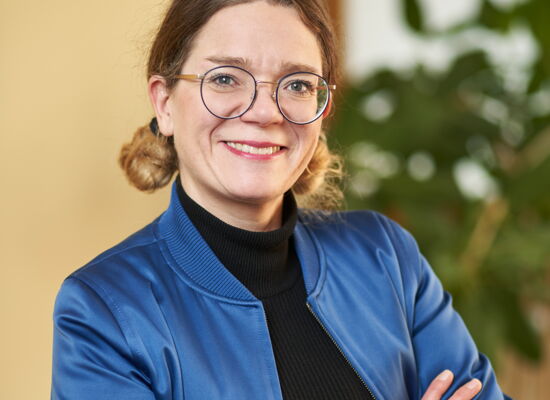 Annika Harder steht lächelnd mit verschränkten Armen vor der Kamera. Sie trägt eine runde Brille, einen schwarzen Rollkragen und eine blaue Jacke.