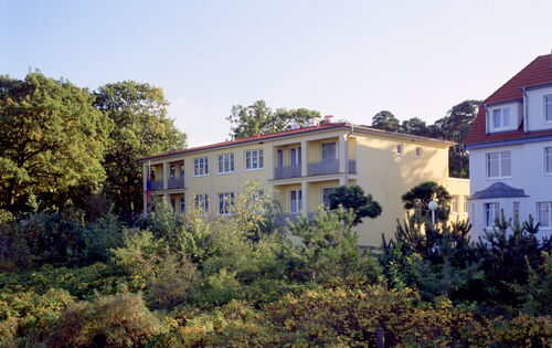 Das AURA-Hotel Boltenhagen, ein gelbes eckiges Gebäude mit schlichten Balkonen und rotem flachen Dach, steht zwischen Büschen und Bäumen. Daneben ist ein weiteres großes, weißes Haus zu sehen.