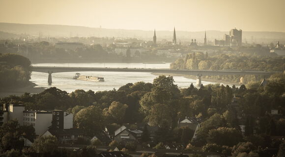 Blick in die Ferne auf die Silhouette von Bonn. Eine lange Brücke spannt sich in der Mitte des Bildes über den Rhein.