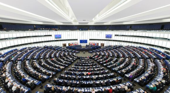 Ein Blick von schräg oben auf das Europäische Parlament in Straßburg: Abgeordnete sitzen  an langen Tischreihen, die ein Halbrund bilden. Strahlenförmig  dazwischen verlaufen Gänge. Auf den Tischen liegen Unterlagen.