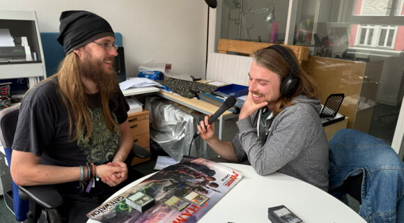 Felix Högl und Christian Balbach sitzen gemeinsam an einem Tisch, auf dem Spielkarten von Magic: The Gathering sowie eine Spielunterlage liegen. Beide sind ins Gespräch vertieft und lachen.